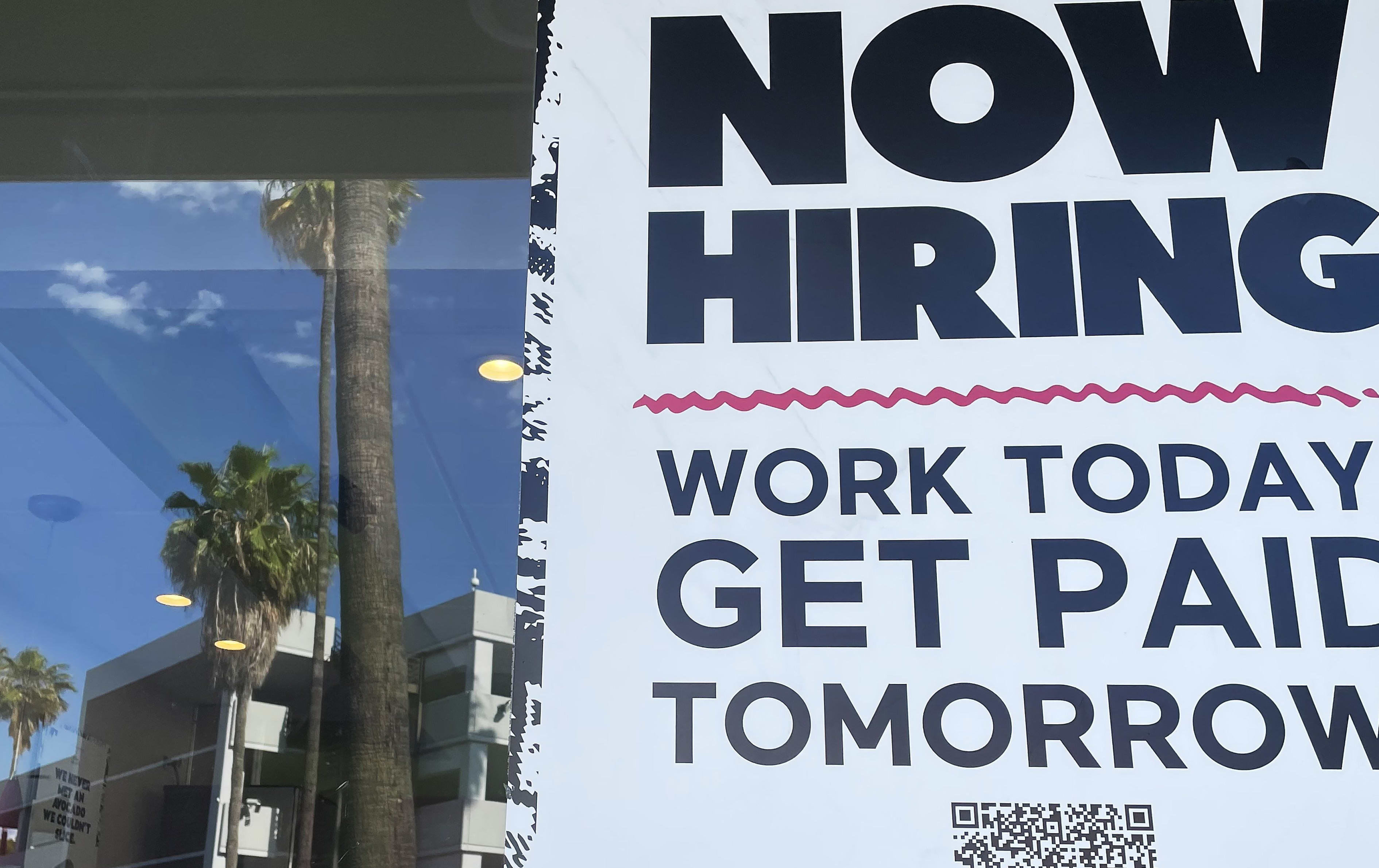 A "Now Hiring" sign on a building with palm trees reflected in the window.