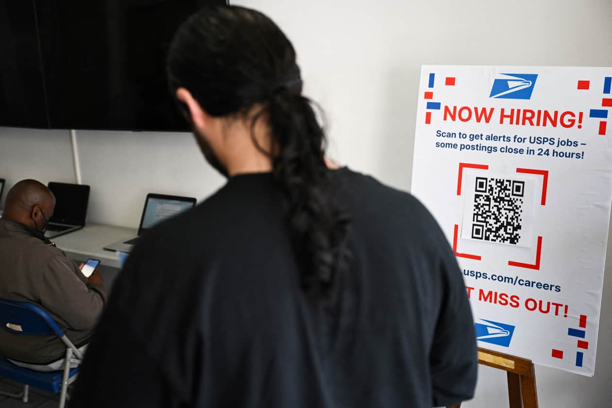 A now hiring sign is displayed as job seekers fill out applications for employment at a United States Postal Service.