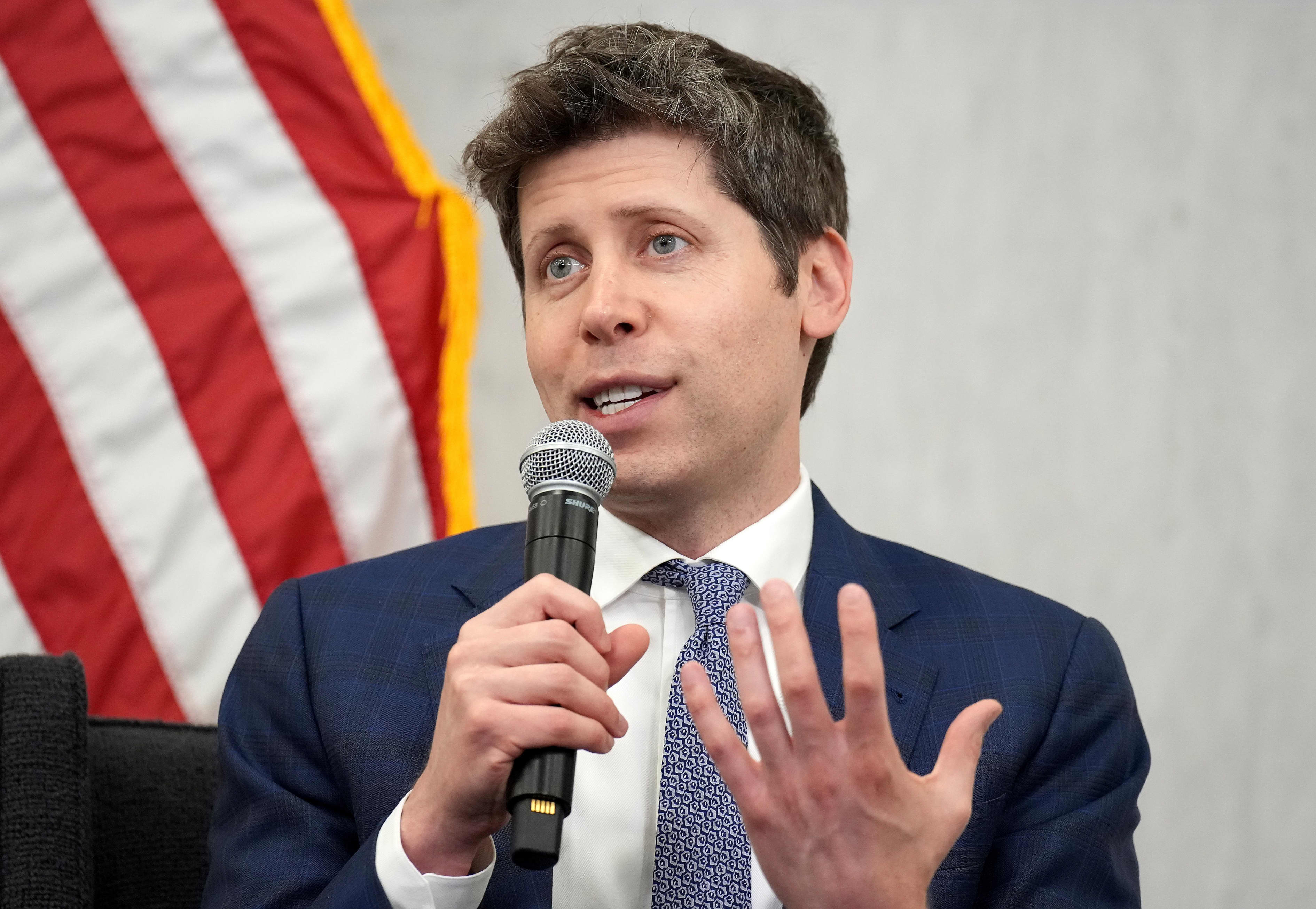 Sam Altman, CEO of OpenAI, speaking into a microphone with an American flag in the background.