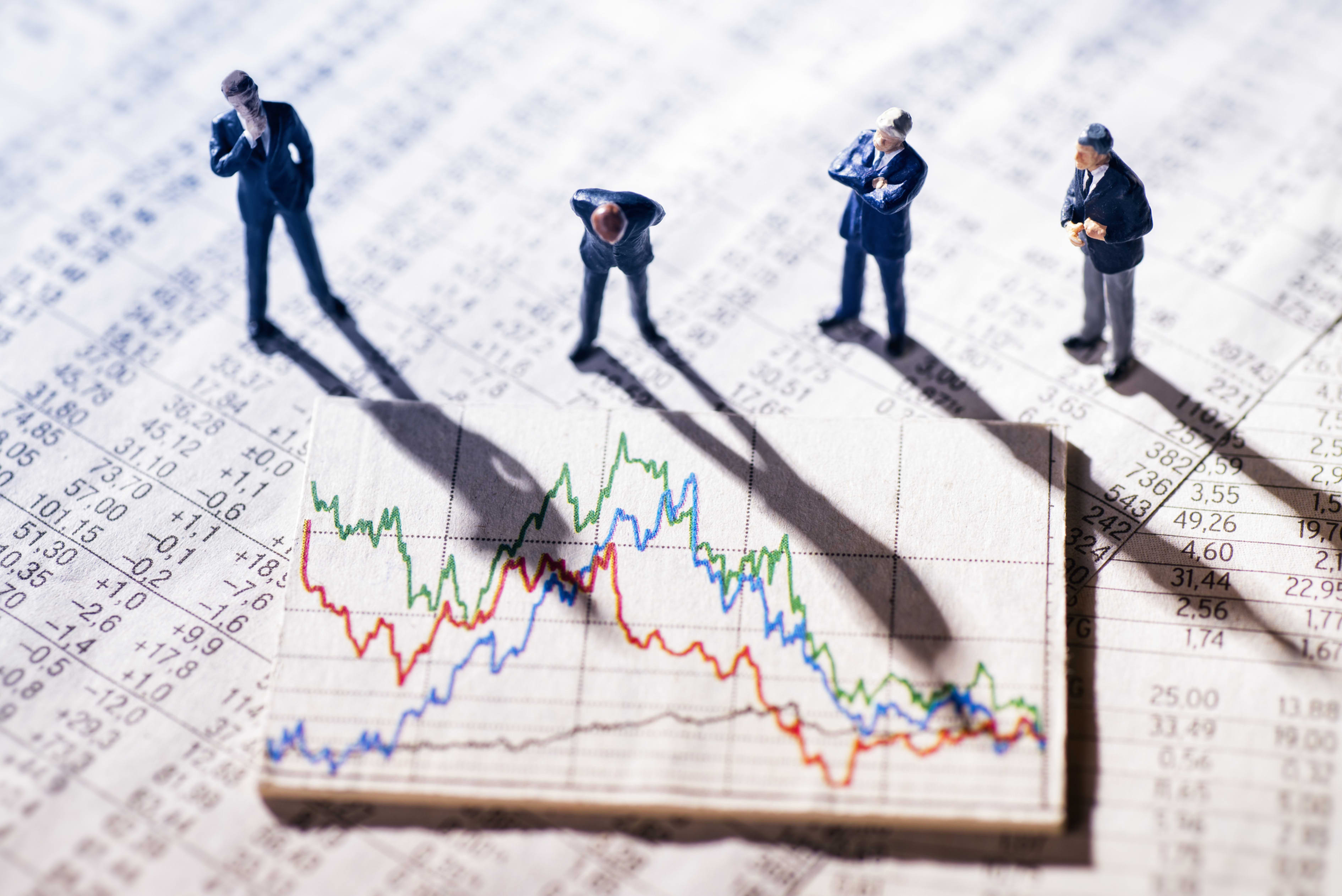 Four miniature business figures examine a newspaper printout of stock market data with a line graph.