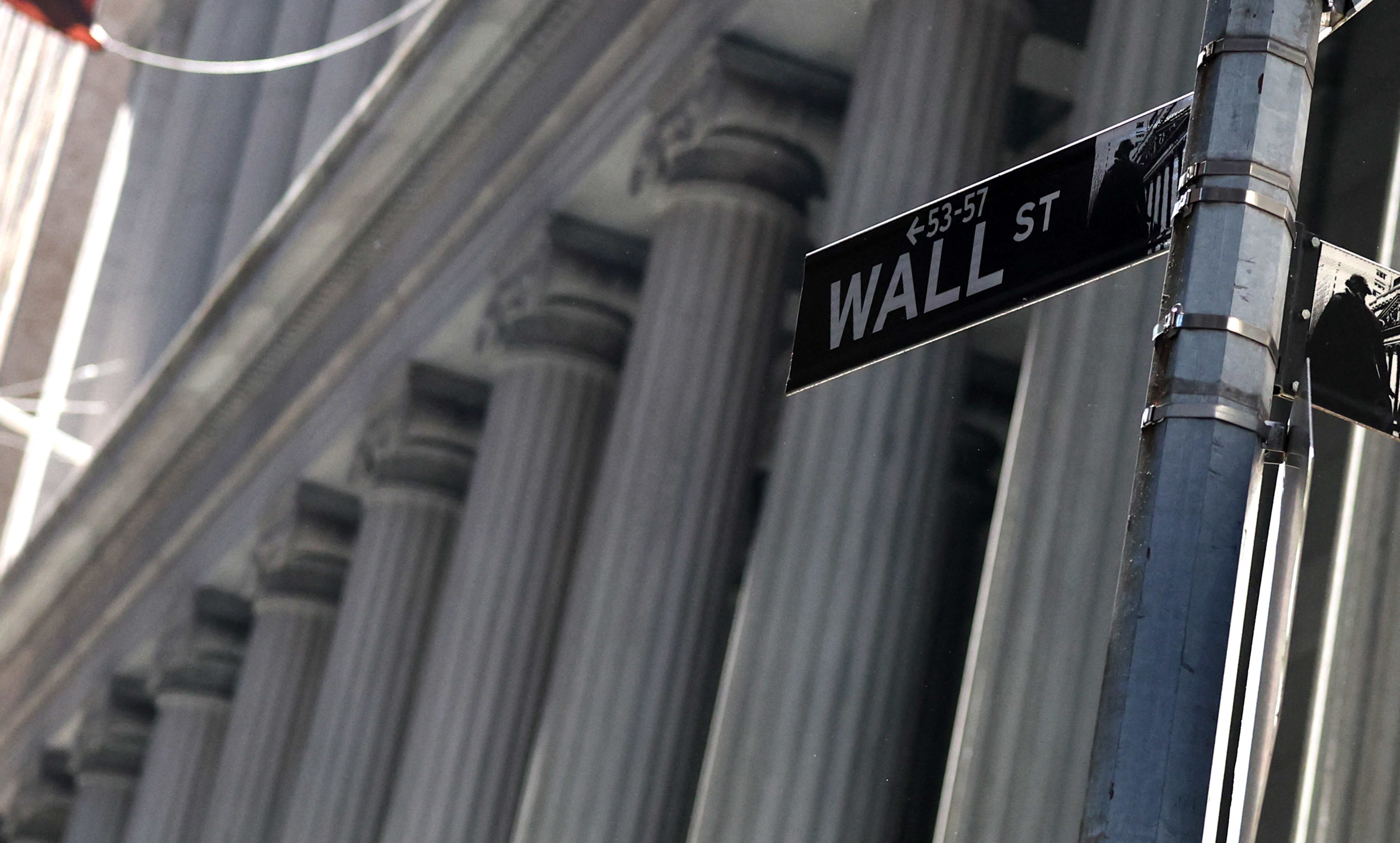 A "Wall St" street sign with the New York Stock Exchange in the background.